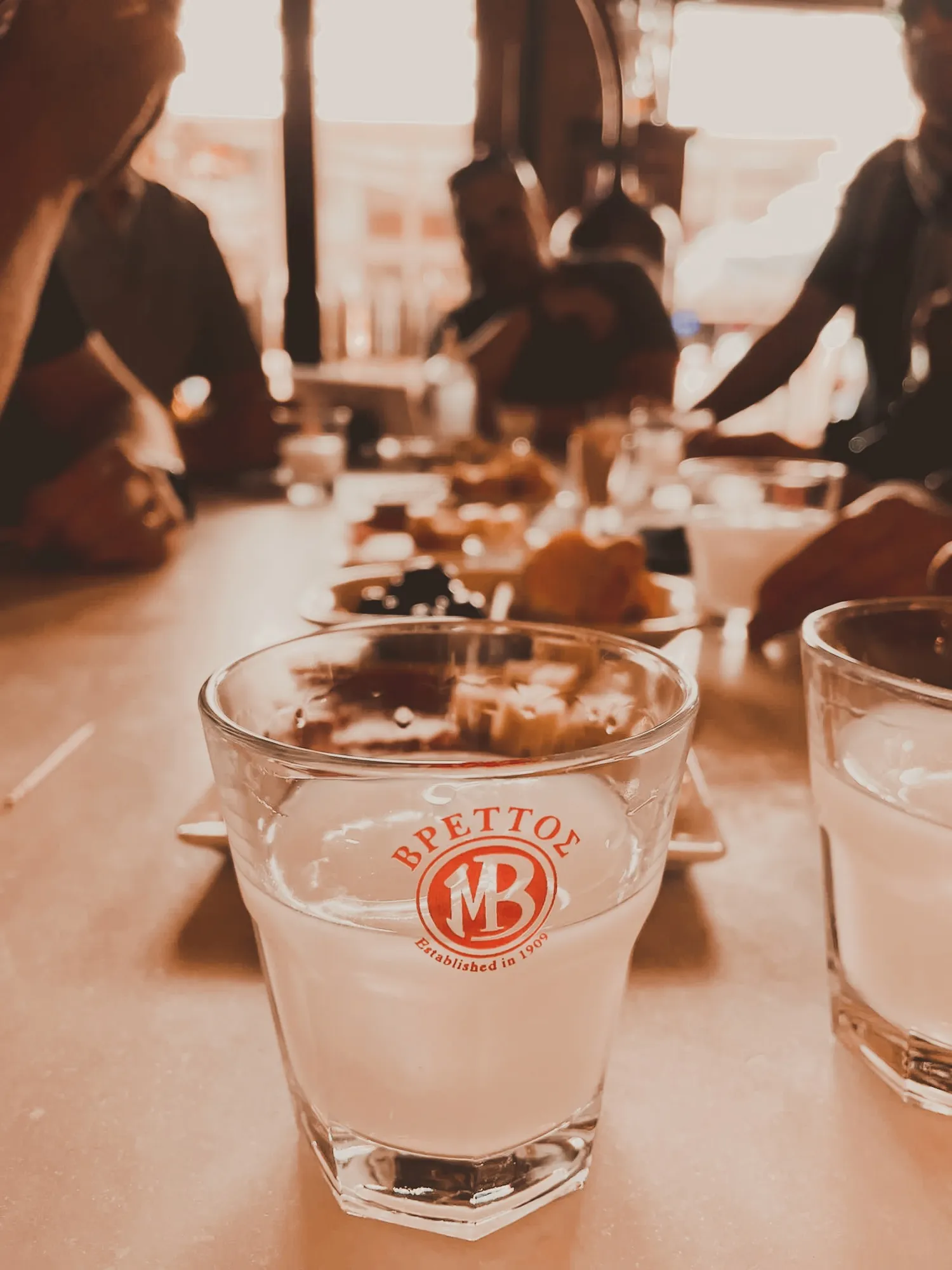 Mehrere Personen sitzen um einen Tisch mit Snacks, im Vordergrund steht ein Glas mit weißlichem Getränk und rotem Brettos Logo